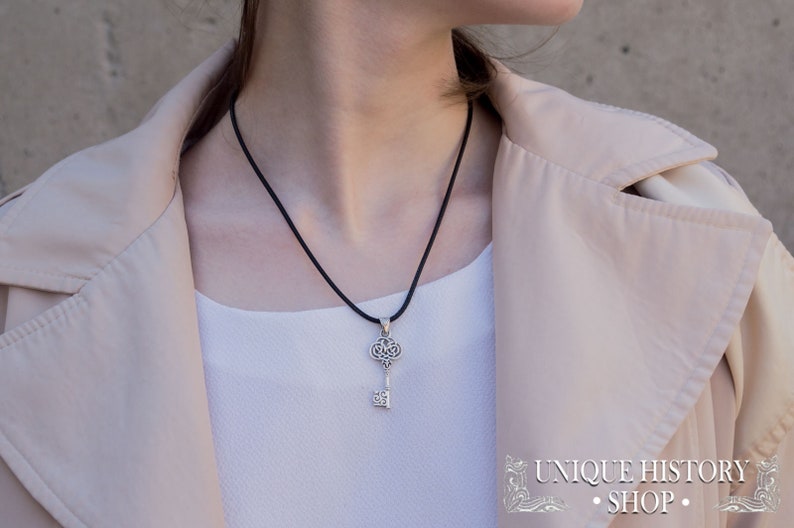 K&ouml;nnte beinhalten: Eine Halskette mit einem silbernen Schl&uuml;sselanh&auml;nger an einem schwarzen Band. Der Schl&uuml;ssel hat ein filigranes Design und ist ein beliebter Stil f&uuml;r Schmuck.