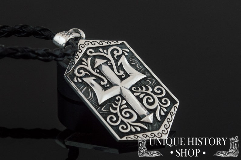 May include: Silver pendant necklace with a detailed trident design surrounded by ornate scrollwork. The pendant is attached to a black leather cord.