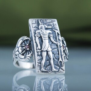 May include: Silver ring with a rectangular design featuring an Egyptian god with a jackal head. The ring has two red gemstones on either side of the central design.