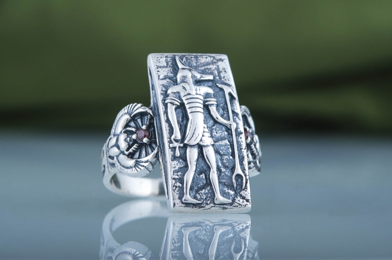 May include: Silver ring with a rectangular design featuring an Egyptian god with a jackal head. The ring has two red gemstones on either side of the central design.
