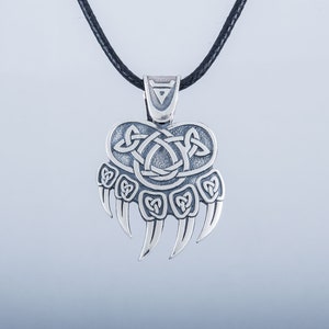 May include: Silver pendant necklace featuring a bear paw print with a Celtic knot design. The pendant is on a black cord.