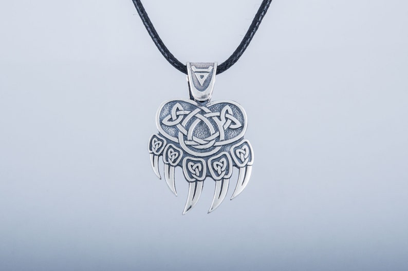 May include: Silver pendant necklace featuring a bear paw print with a Celtic knot design. The pendant is on a black cord.