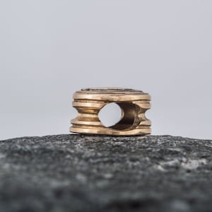 May include: A gold-colored metal bead with a circular hole in the center. The bead has a textured surface and is sitting on a gray rock.