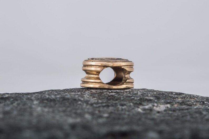 May include: A gold-colored metal bead with a circular hole in the center. The bead has a textured surface and is sitting on a gray rock.