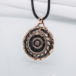 Pode incluir: Um colar com um pingente redondo com um design de drag&atilde;o preto e uma pequena gema rosa no centro. O pingente &eacute; feito de metal e tem um acabamento em ouro rosa.