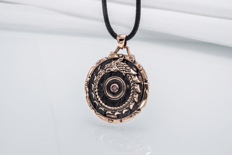 Pode incluir: Um colar com um pingente redondo com um design de drag&atilde;o preto e uma pequena gema rosa no centro. O pingente &eacute; feito de metal e tem um acabamento em ouro rosa.