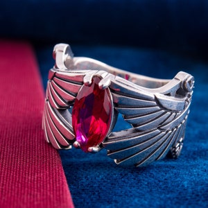 May include: A silver ring with a marquise-cut, ruby-red gemstone. The ring has wing-like designs on either side of the gemstone. The ring is shown on a blue velvet surface with a red fabric edge.