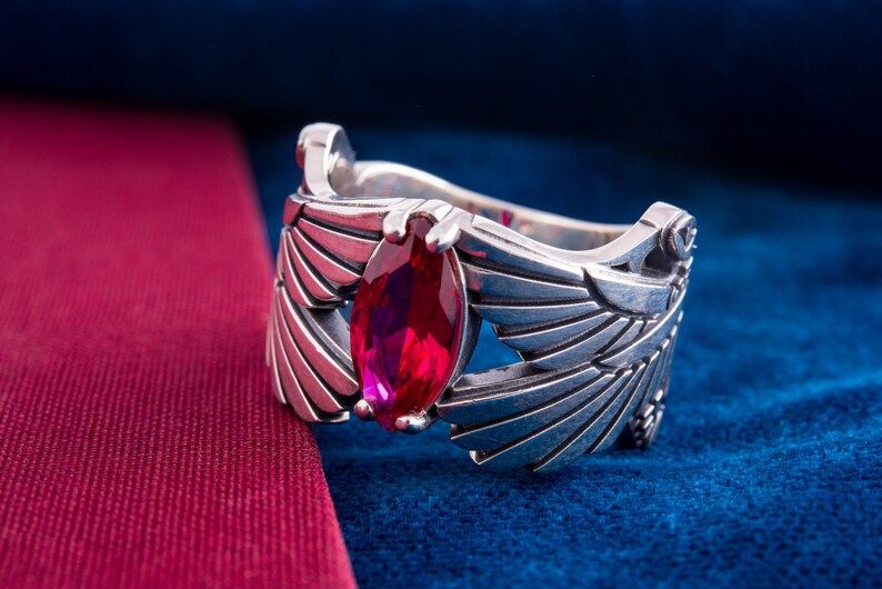 May include: A silver ring with a marquise-cut, ruby-red gemstone. The ring has wing-like designs on either side of the gemstone. The ring is shown on a blue velvet surface with a red fabric edge.
