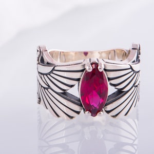 May include: A silver ring with a marquise-cut, deep red gemstone. The ring's band is decorated with stylized wing designs. The gemstone is centrally set, providing a focal point to the jewellery.