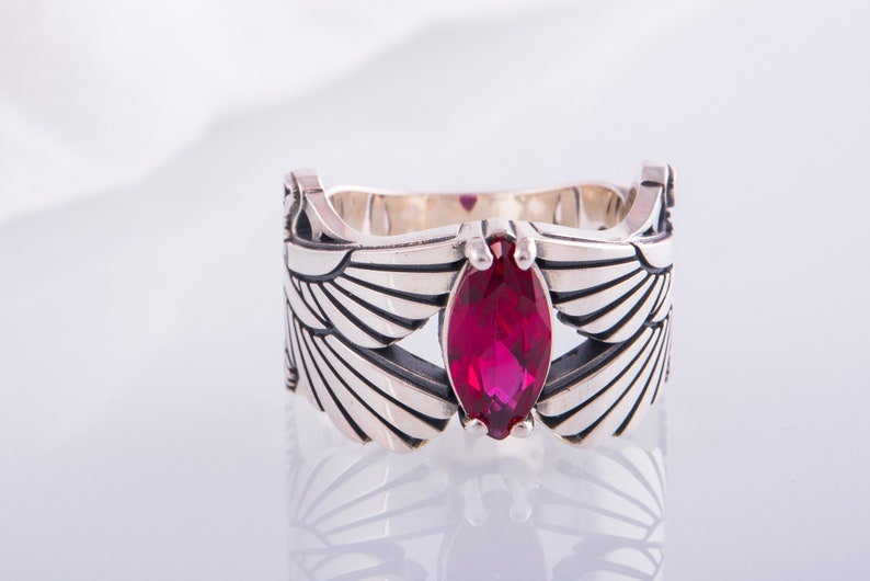 May include: A silver ring with a marquise-cut, deep red gemstone. The ring's band is decorated with stylized wing designs. The gemstone is centrally set, providing a focal point to the jewellery.