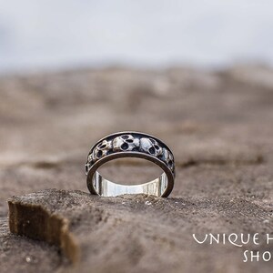 May include: A silver ring with a band of skulls around the circumference. The ring is sitting on a piece of wood.