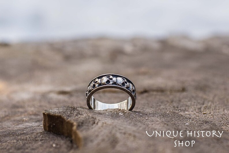 May include: A silver ring with a band of skulls around the circumference. The ring is sitting on a piece of wood.