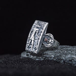 May include: A silver ring with an engraved image of an Egyptian god and a scarab beetle with a red stone. The ring is on a gray rock.