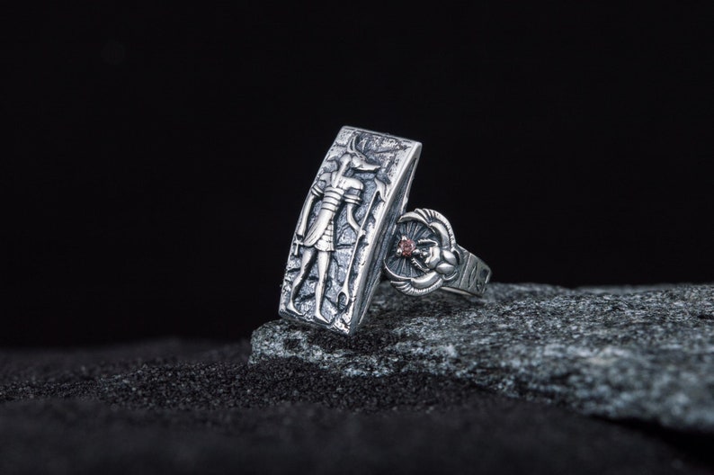 May include: A silver ring with an engraved image of an Egyptian god and a scarab beetle with a red stone. The ring is on a gray rock.