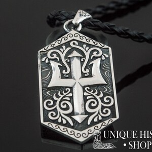 May include: Silver pendant necklace with a detailed design of a trident and floral patterns. The pendant is shaped like a shield and has a black leather cord.