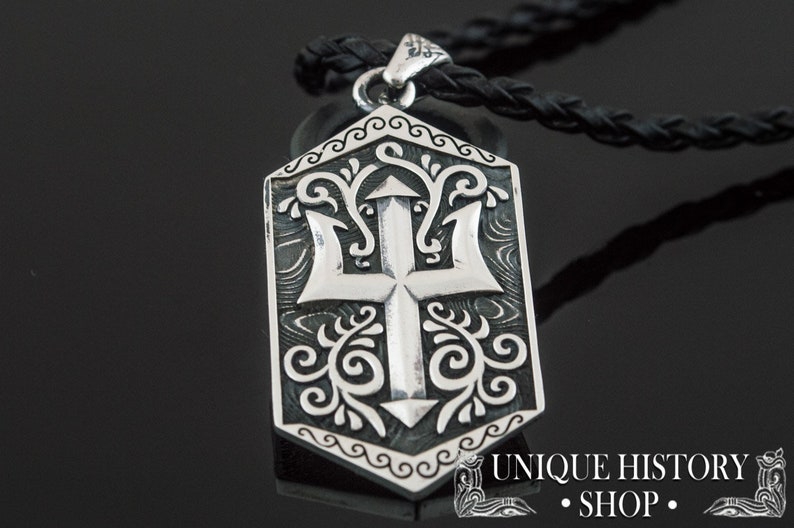 May include: Silver pendant necklace with a detailed design of a trident and floral patterns. The pendant is shaped like a shield and has a black leather cord.