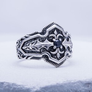 May include: A silver-colored ring with an ornate design. The ring features a central black gemstone and detailed scrollwork. The design includes a fleur-de-lis motif. The ring is set against a white background.
