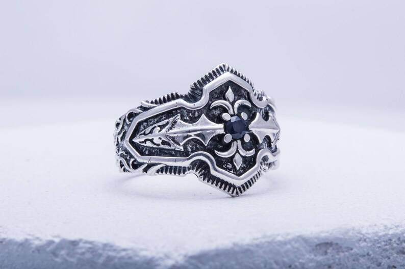May include: A silver-colored ring with an ornate design. The ring features a central black gemstone and detailed scrollwork. The design includes a fleur-de-lis motif. The ring is set against a white background.