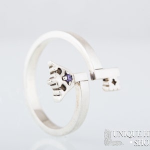 May include: A silver ring with a key design. The key has a purple gemstone in the center. The ring is open and the key is movable.