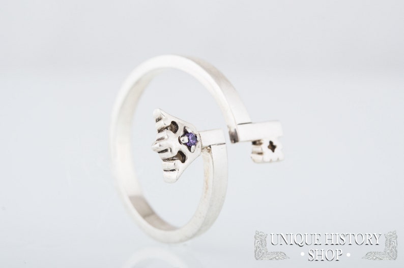 May include: A silver ring with a key design. The key has a purple gemstone in the center. The ring is open and the key is movable.