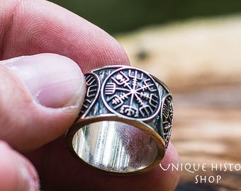May include: A silver-colored ring with a detailed Viking design. The ring features a central symbol surrounded by intricate patterns. The ring is held in a hand, with the text "UNIQUE HISTORY SHOP" visible in the background.