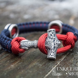 Puede incluir: Un brazalete trenzado rojo y azul con un amuleto Mjolnir de plata. El amuleto est&aacute; detallado con intrincados nudos y un dise&ntilde;o en relieve. El brazalete est&aacute; atado con un nudo y tiene un anillo de plata en cada extremo.