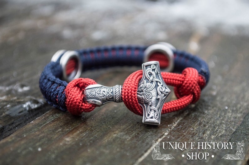 Puede incluir: Un brazalete trenzado rojo y azul con un amuleto Mjolnir de plata. El amuleto est&aacute; detallado con intrincados nudos y un dise&ntilde;o en relieve. El brazalete est&aacute; atado con un nudo y tiene un anillo de plata en cada extremo.