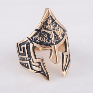 Pode incluir: Um anel dourado com um design detalhado de um capacete espartano. O capacete &eacute; texturizado e tem um padr&atilde;o de chave grega ao redor da banda.