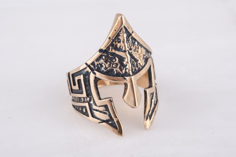 Pode incluir: Um anel dourado com um design detalhado de um capacete espartano. O capacete &eacute; texturizado e tem um padr&atilde;o de chave grega ao redor da banda.
