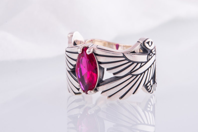 May include: A silver ring with a marquise-cut, bright pink gemstone. The ring's design includes stylised wings on either side of the gemstone, with detailed feather etching. The ring is set against a white background.