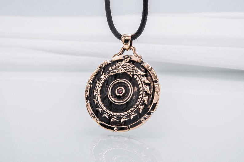 Pode incluir: Um pingente redondo de bronze com um design de drag&atilde;o preto e uma pedra preciosa vermelha no centro. O pingente est&aacute; preso a um cord&atilde;o de couro preto.