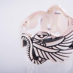 May include: A silver ring featuring an elaborate wing motif and a rectangular red gemstone. The ring is intricately detailed, with etched lines and curves. The gemstone is set into the ring's band, providing a vibrant contrast.