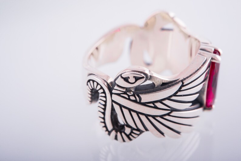May include: A silver ring featuring an elaborate wing motif and a rectangular red gemstone. The ring is intricately detailed, with etched lines and curves. The gemstone is set into the ring's band, providing a vibrant contrast.