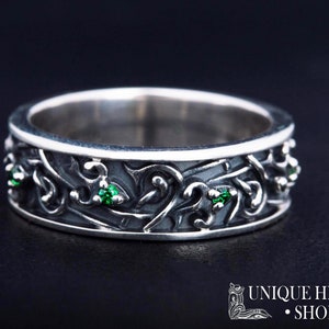 May include: A silver ring with an intricate design featuring green gemstones. The ring is wide and has a textured surface.