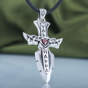 May include: Silver pendant necklace featuring a sword-shaped cross with a red gemstone at the center. The sword is inscribed with the words "Valhalla" in an ancient script.