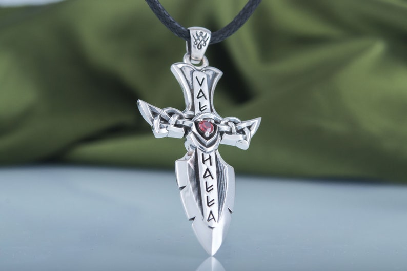 May include: Silver pendant necklace featuring a sword-shaped cross with a red gemstone at the center. The sword is inscribed with the words "Valhalla" in an ancient script.