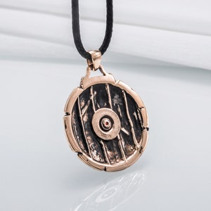 Pode incluir: Um pingente redondo de bronze com um design detalhado de um escudo. O escudo tem um ponto vermelho central e &eacute; rodeado por uma borda elevada.