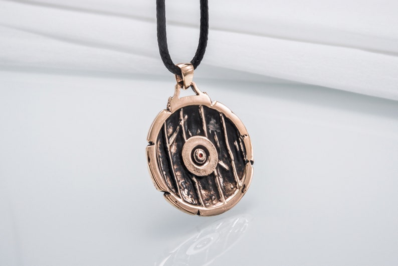 Pode incluir: Um pingente redondo de bronze com um design detalhado de um escudo. O escudo tem um ponto vermelho central e &eacute; rodeado por uma borda elevada.