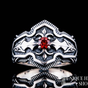 May include: A silver ring with an ornate design featuring a central red gemstone. The ring has a detailed, textured pattern with a dark background. The ring is likely made of sterling silver.