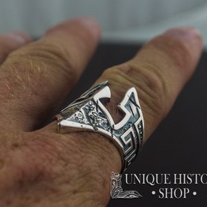 Pode incluir: Um anel de prata com um design detalhado de um capacete com um padr&atilde;o de chave grega. O anel &eacute; usado no dedo de uma pessoa.