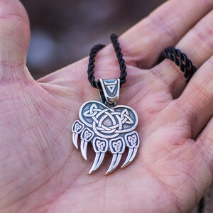 May include: A silver bear paw pendant with a Celtic knot design. The pendant is on a black cord necklace.
