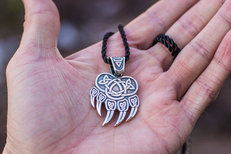 May include: A silver bear paw pendant with a Celtic knot design. The pendant is on a black cord necklace.
