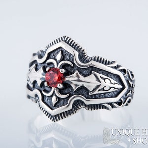 May include: A silver ring with an ornate design featuring a central red gemstone. The ring has a detailed pattern with arrow and floral motifs. The ring is from the Unique History Shop.