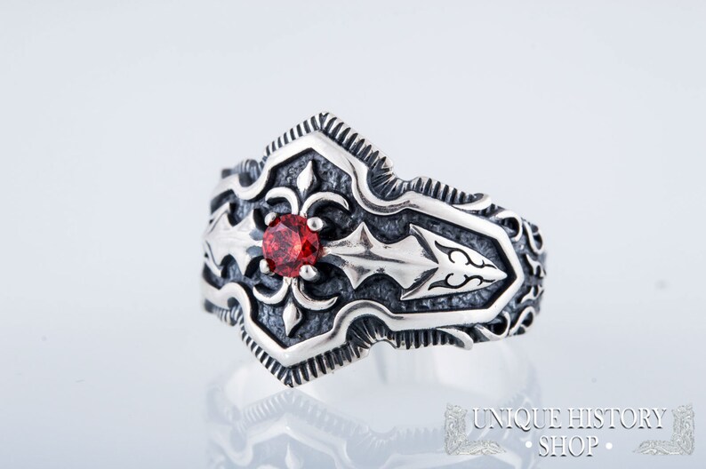 May include: A silver ring with an ornate design featuring a central red gemstone. The ring has a detailed pattern with arrow and floral motifs. The ring is from the Unique History Shop.
