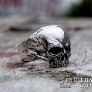 Pu&ograve; includere: Un anello in argento a forma di teschio con dettagli intricati. Il teschio &egrave; rivolto in avanti con un viso e denti dettagliati. La fascia dell'anello &egrave; larga e presenta un design testurizzato.