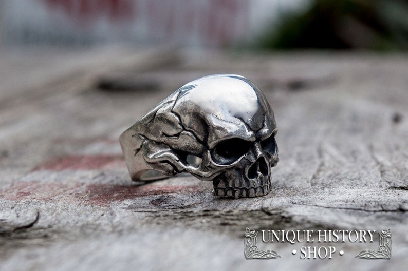 Pu&ograve; includere: Un anello in argento a forma di teschio con dettagli intricati. Il teschio &egrave; rivolto in avanti con un viso e denti dettagliati. La fascia dell'anello &egrave; larga e presenta un design testurizzato.
