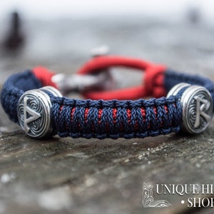 Puede incluir: Una pulsera trenzada azul y roja con dos abalorios plateados que muestran un s&iacute;mbolo vikingo. La pulsera est&aacute; sobre una superficie de madera.
