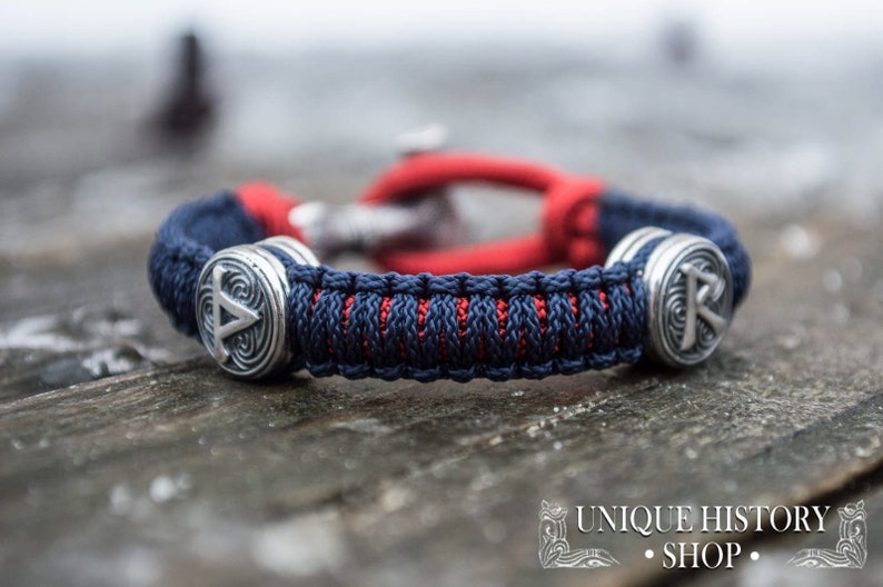 Puede incluir: Una pulsera trenzada azul y roja con dos abalorios plateados que muestran un s&iacute;mbolo vikingo. La pulsera est&aacute; sobre una superficie de madera.