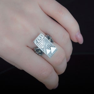 May include: A silver ring with a rectangular design featuring a stylized mountain scene and geometric patterns. The ring has a textured surface and a floral design on the side.