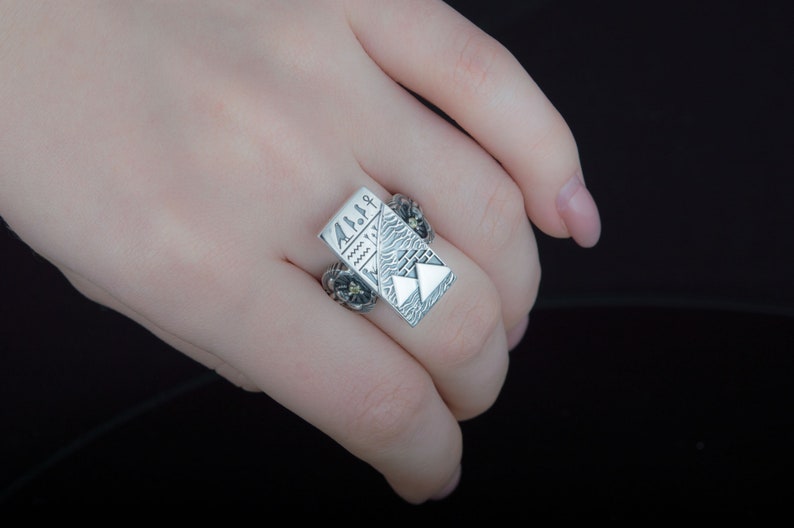 May include: A silver ring with a rectangular design featuring a stylized mountain scene and geometric patterns. The ring has a textured surface and a floral design on the side.